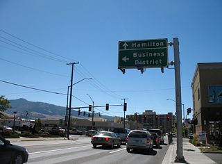 An overhead sign assembly precedes intersection between Orange and Broadway Streets (Business Loop I-90) on U.S. 93 Business south. The guide sign alludes to Orange Street as the main line for U.S. 93, directing motorists to Hamilton, the U.S. 93 destination south of Missoula in 47 miles.